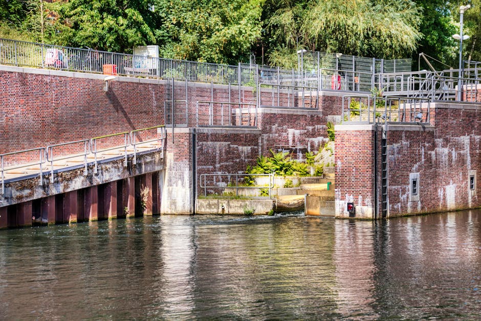 View of a brick riverbank retaining wall with metal railings and steps leading down to the water, situated near Teddington Lock. The wall features multiple sections with varying heights, some with sma