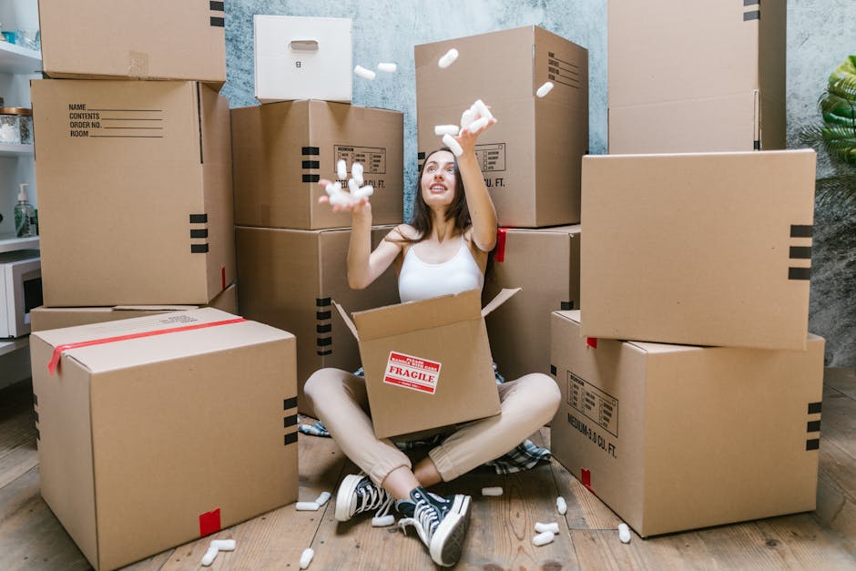 A young woman sitting cross-legged on a wooden floor surrounded by multiple cardboard boxes of various sizes, some sealed with red tape and others labeled with black markings, while she is cheerfully 